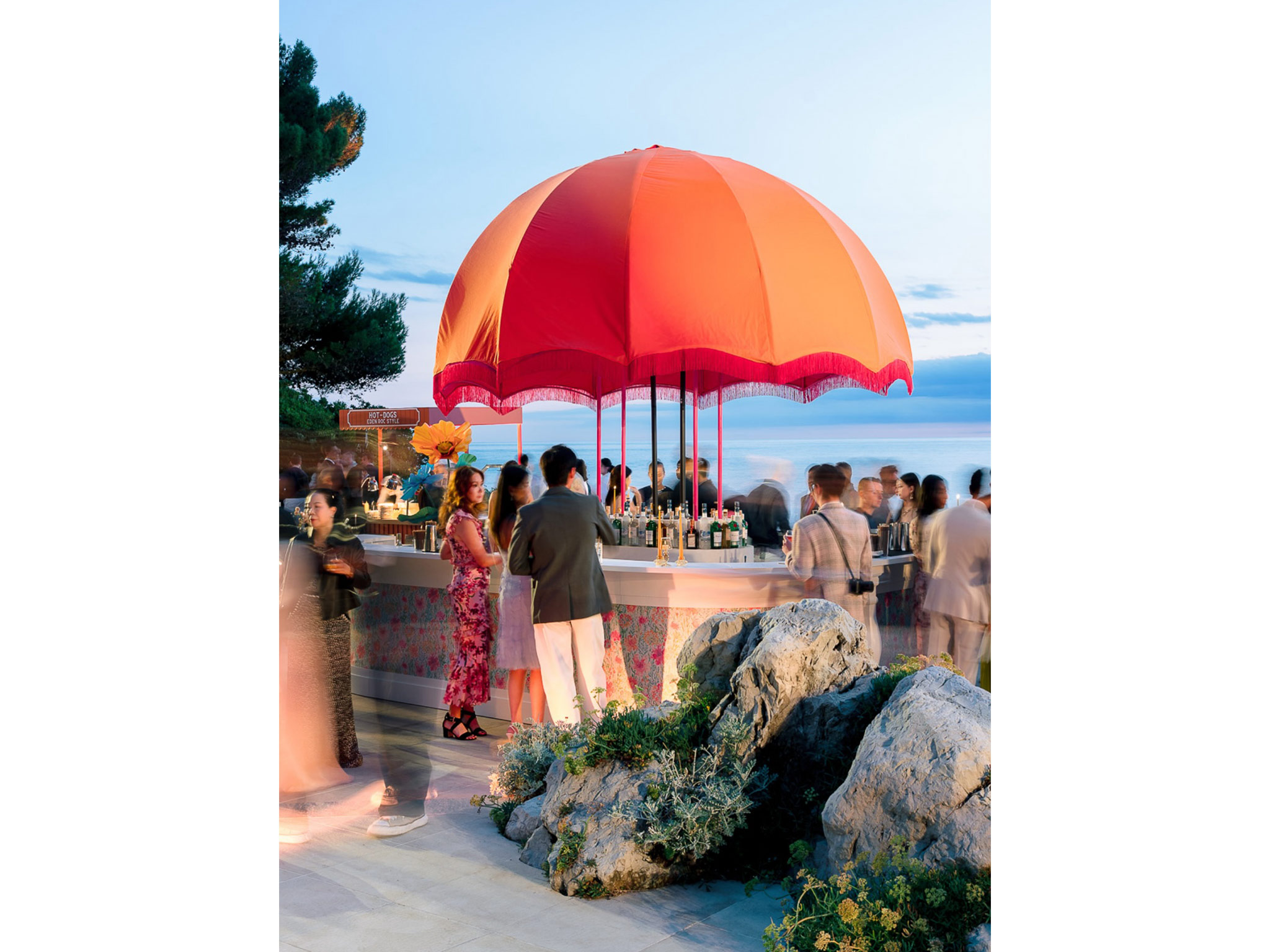 Vue d'ensemble du bar central arrondi avec dôme orangé à franges et grandes fleurs géantes décoratives, entouré de convives lors du cocktail du Welcome Dinner d'Angelina & Kelvin — production AUCOP à l'Hôtel du Cap Eden Roc, vue sur la Méditerranée.