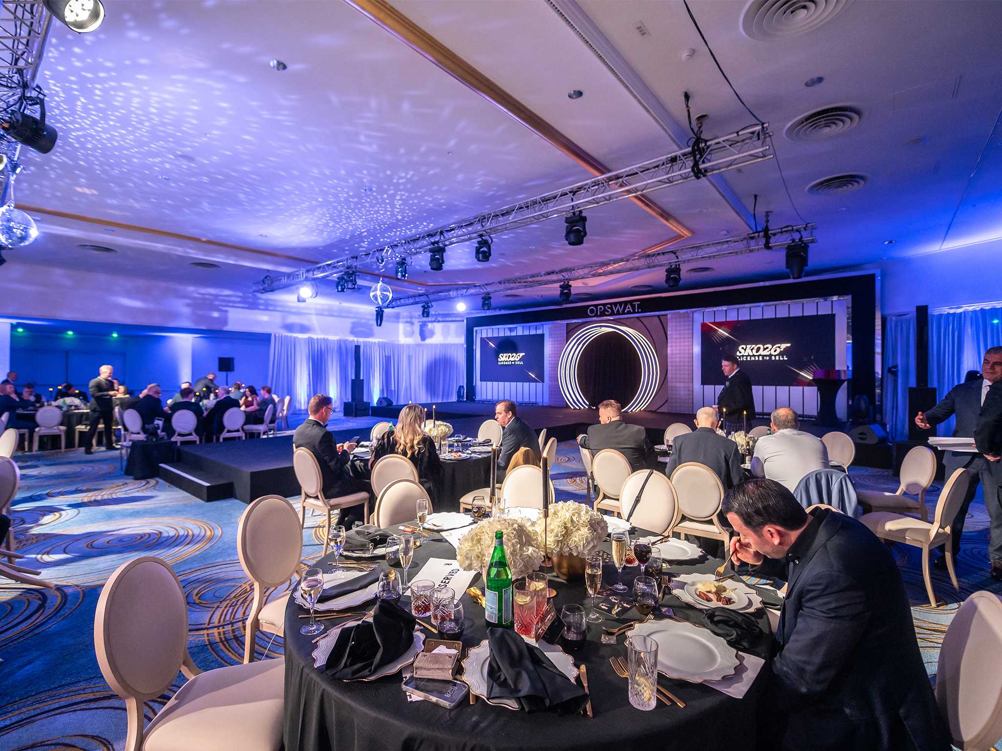 Soirée de gala du SKO26 d'OPSWAT au Fairmont Monaco mise en scène par AUCOP : tables rondes dressées sous un éclairage bleu et boules à facettes, face à la scène signée AUCOP — arches à LED incrustées façon canon de pistolet, backdrop sur mesure et bandeau OPSWAT, encadrés de deux murs LED ROE Diamond 2.6 mm diffusant « SKO26 – License to Sell ».
