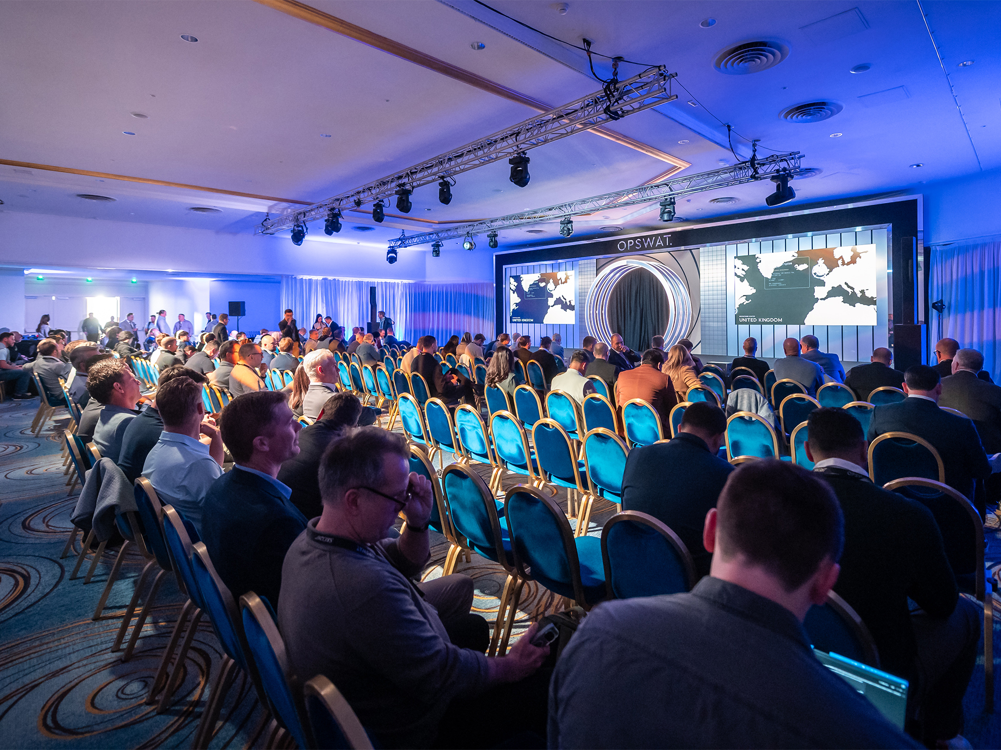 Session du SKO26 d'OPSWAT au Fairmont Monaco : la scénographie AUCOP — arche centrale à LED, backdrop sur mesure et bandeau OPSWAT — accueille une présentation « United Kingdom » sur les deux murs LED ROE Diamond 2.6 mm, dans une ambiance lumineuse bleue et une diffusion sonore signées AUCOP.