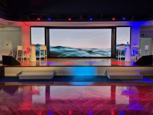 Scène de la salle plénière à bord du Club Med 2, port de Nice : mur LED grand format diffusant une image de vagues, éclairage scénique rouge et bleu, retours de scène, parquet — installation technique AUCOP pour la convention corporate