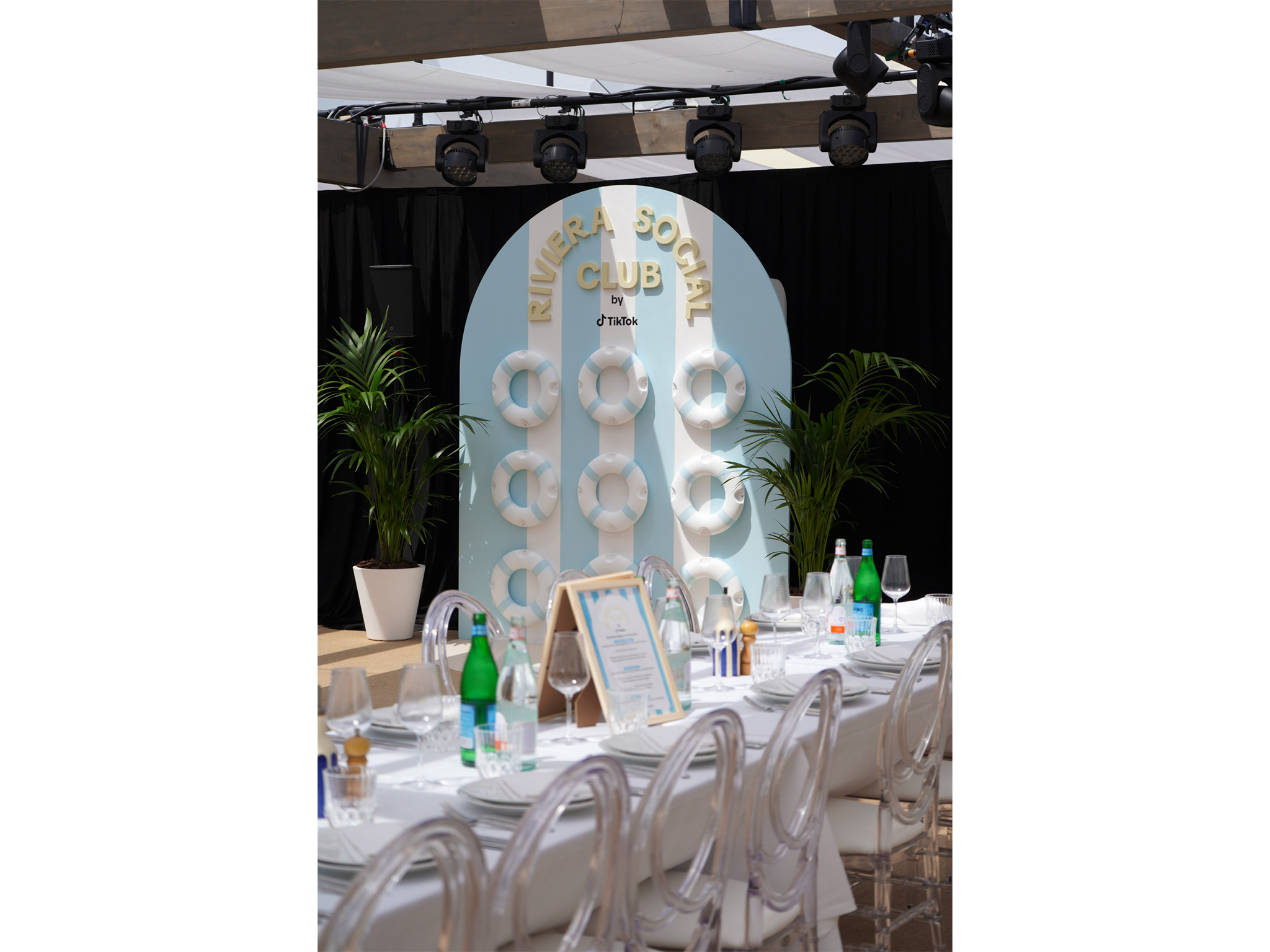 Structure de photocall en arche arrondie créée par AUCOP pour le Riviera Social Club by TikTok : fond à rayures blanches et bleu ciel décoré de six bouées de sauvetage rétro et entouré de plantes vertes en pots blancs.