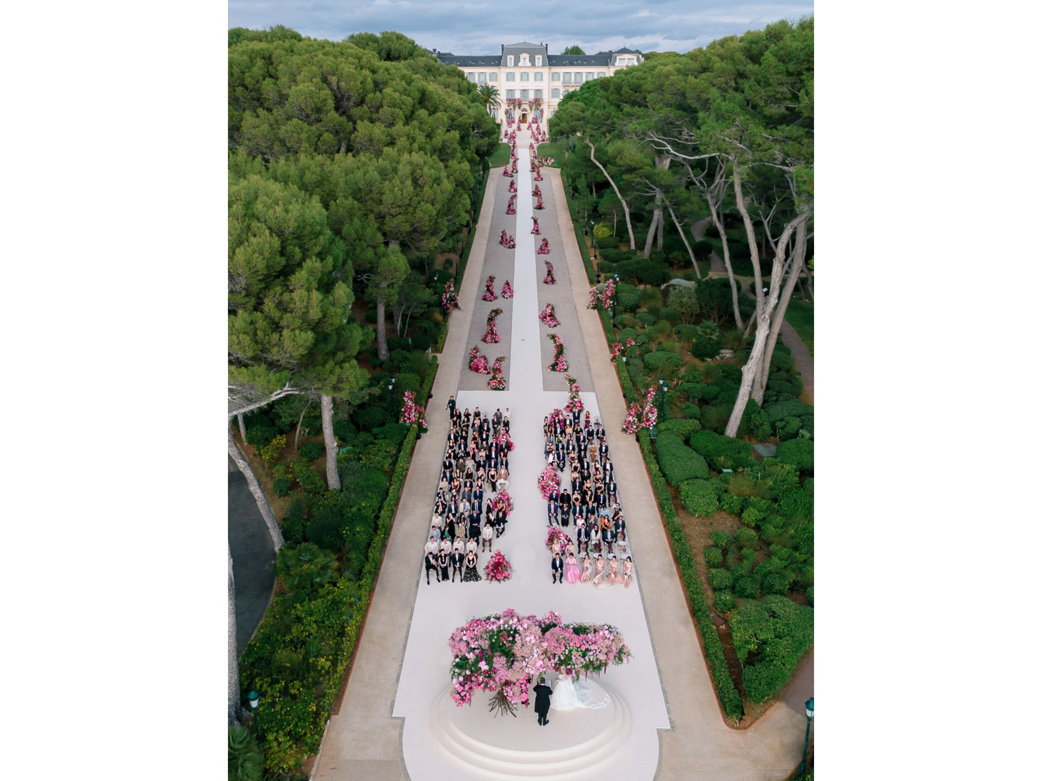 Vue aérienne de la cérémonie de mariage à l'Hôtel du Cap-Eden-Roc : tapis blanc, colonnes florales roses, invités, château en arrière-plan — production AUCOP