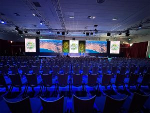Salle d'une plénière corporate au Club Med d'Opio en phase de préparation — rangées de chaises, écrans de rappel latéraux et éclairage bleu AUCOP