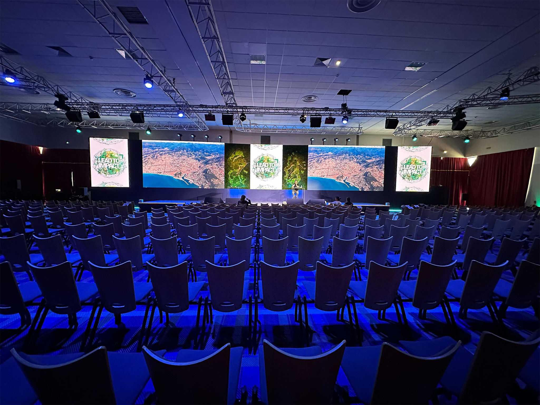 Salle d'une plénière corporate au Club Med d'Opio en phase de préparation — rangées de chaises, écrans de rappel latéraux et éclairage bleu AUCOP