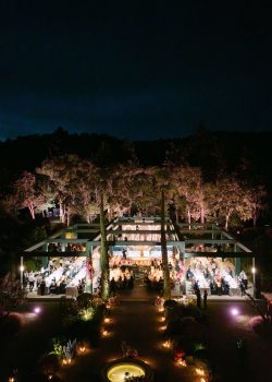 AUCOP - Vue aérienne au crépuscule du dîner de mariage : pergola-structure verte illuminée, allée en gravier avec fontaines fleuries et stèles florales, jardin provençal - scénographie White Eden Weddings, production AUCOP, Airelles Château d'Estoublon, Provence