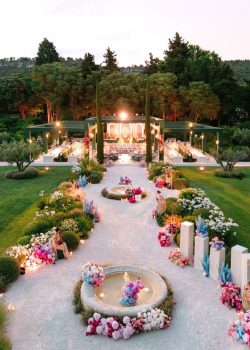 AUCOP - Vue aérienne au crépuscule du dîner de mariage : pergola-structure verte illuminée, allée en gravier avec fontaines fleuries et stèles florales, jardin provençal - scénographie White Eden Weddings, production AUCOP, Airelles Château d'Estoublon, Provence