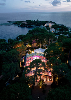 aucop-drone-tente-receptive-bord-de-mer-hotel-cap-eden-roc-nuit-mariage-angelina-kelvin-2026 Vue aérienne de nuit de la tente réceptive illuminée en rose nichée dans les pins au bord de la Méditerranée — mariage Angelina & Kelvin, Hôtel du Cap-Eden-Roc, production audiovisuelle AUCOP