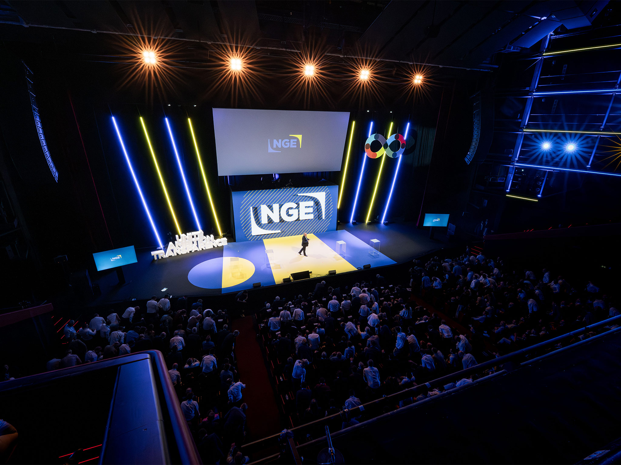 Vue en plongée sur la scène NGE au Palais des Festivals de Cannes : plateau circulaire bleu et jaune, colonnes néon et mise en lumière conçus par AUCOP, intervenant en scène devant plus de 1 000 participants.