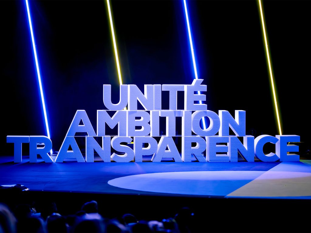 Logo 3D monumental "Unité – Ambition – Transparence" fabriqué sur-mesure par AUCOP, posé sur la scène de l'auditorium du Palais des Festivals de Cannes, baigné dans une lumière bleue avec faisceaux néon.