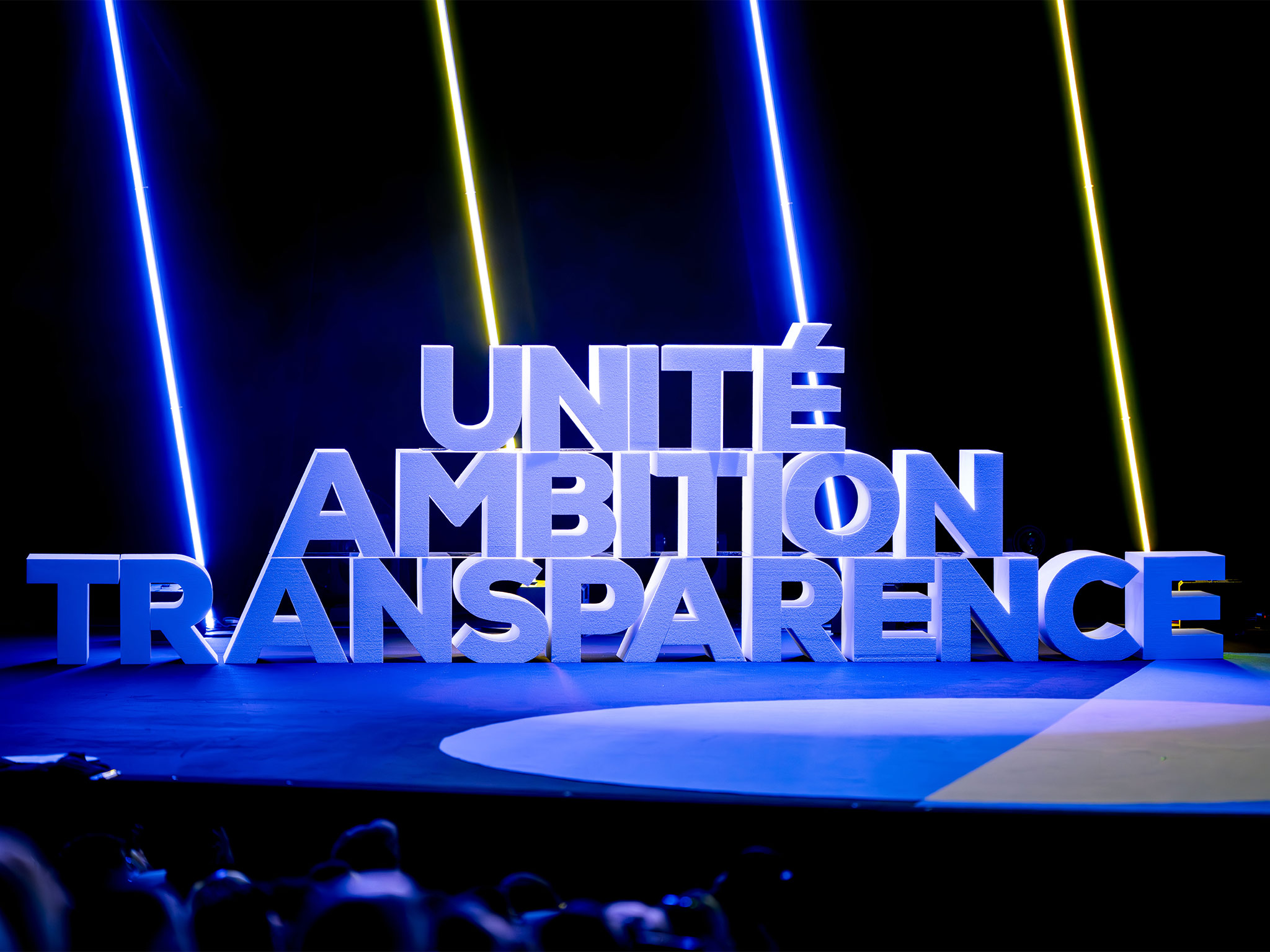 Logo 3D monumental "Unité – Ambition – Transparence" fabriqué sur-mesure par AUCOP, posé sur la scène de l'auditorium du Palais des Festivals de Cannes, baigné dans une lumière bleue avec faisceaux néon.