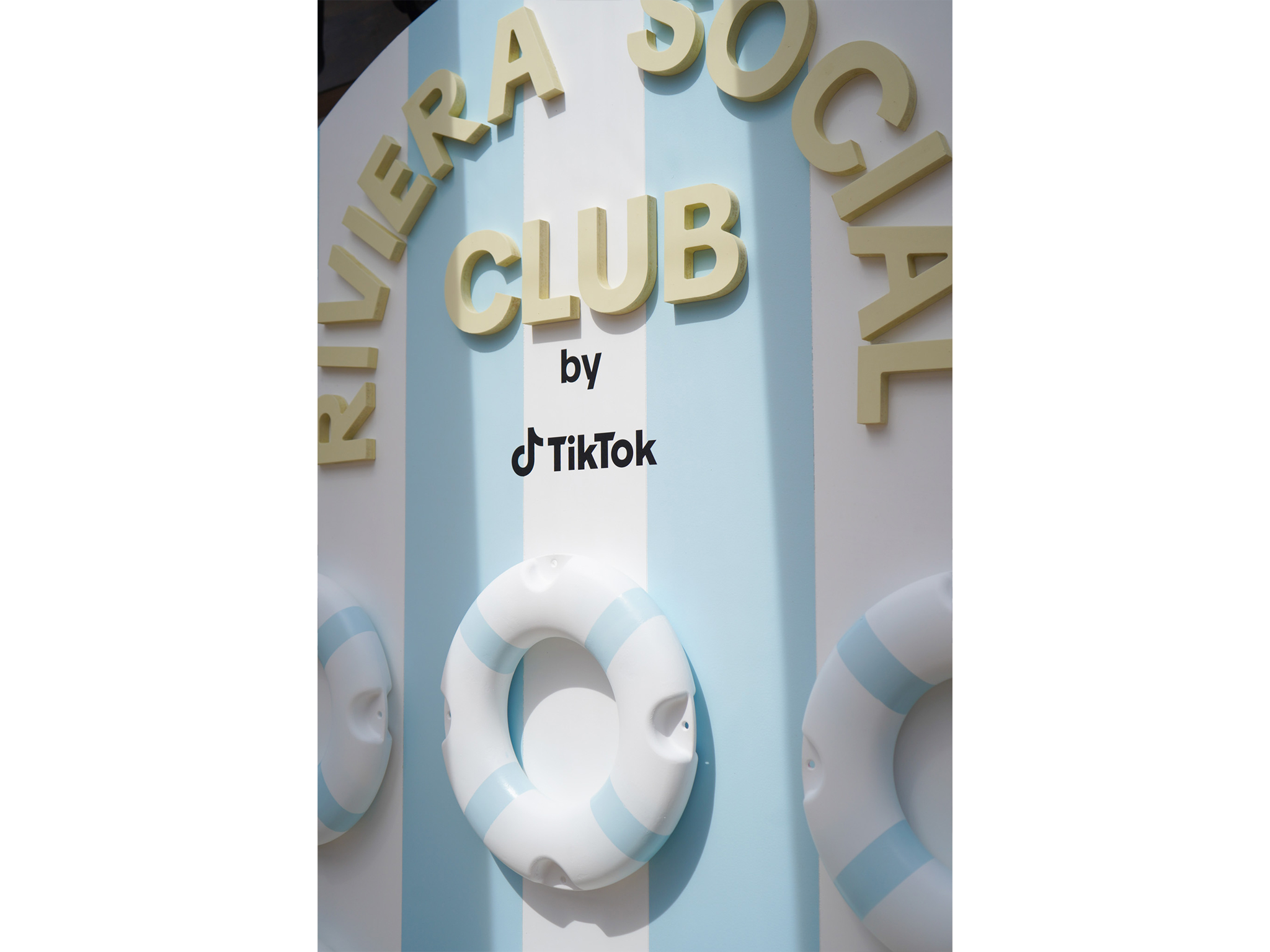 Détail de la signalétique en relief du photocall Riviera Social Club by TikTok à Cannes : lettres jaunes 3D et logo TikTok noir fixés sur un panneau à rayures, au-dessus de bouées de plage décoratives blanches et bleues.