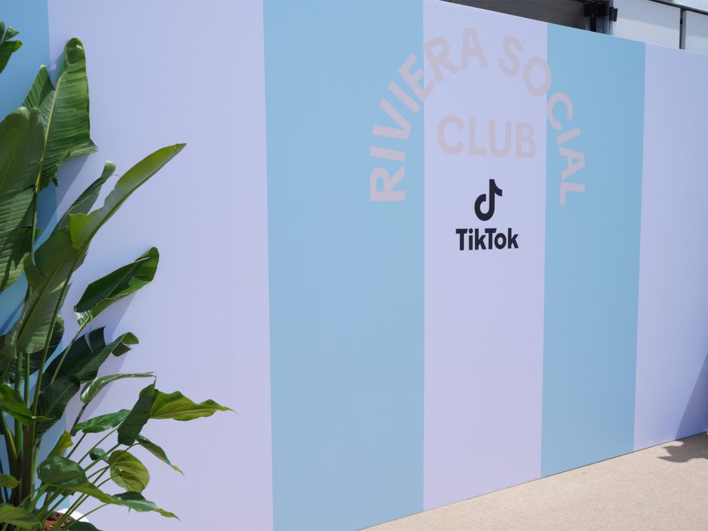 Vue en angle du photocall linéaire Riviera Social Club by TikTok conçu par AUCOP : bâche tendue imprimée sur-mesure à rayures pastel, accompagnée de plantes tropicales pour habiller l'espace éphémère cannois