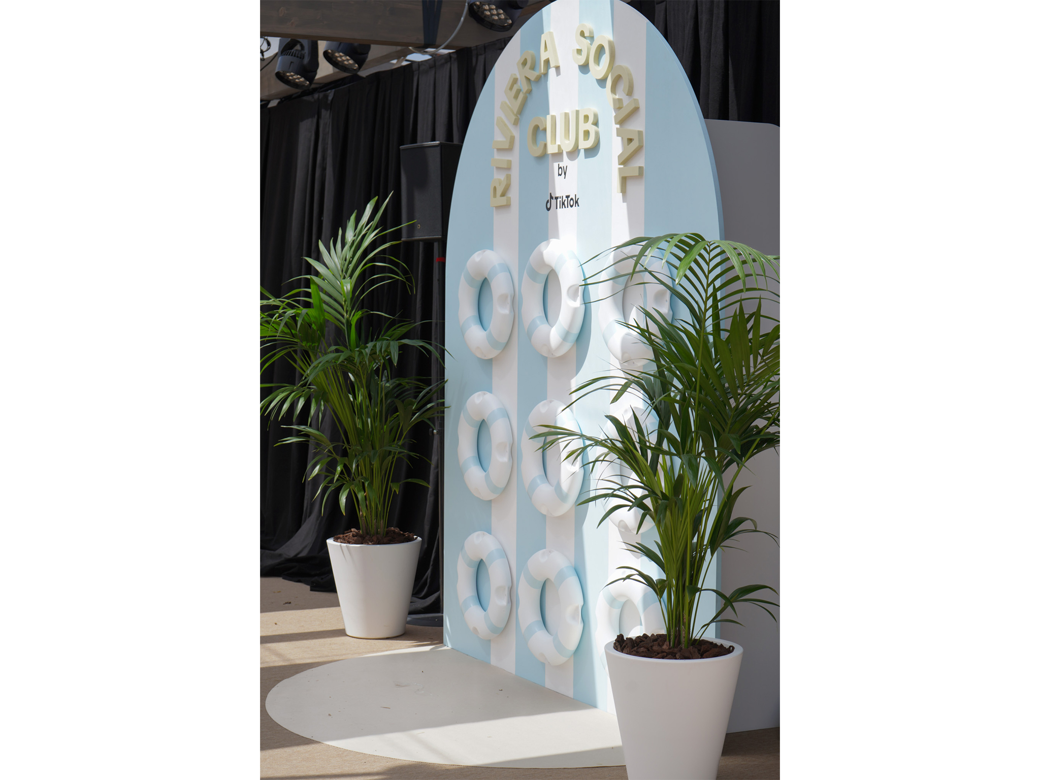 Structure de photocall en arche arrondie créée par AUCOP pour le Riviera Social Club by TikTok : fond à rayures blanches et bleu ciel décoré de six bouées de sauvetage rétro et entouré de plantes vertes en pots blancs.