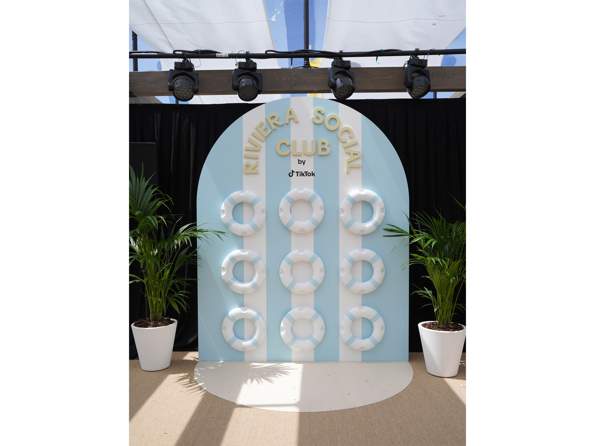 Structure de photocall en arche arrondie créée par AUCOP pour le Riviera Social Club by TikTok : fond à rayures blanches et bleu ciel décoré de six bouées de sauvetage rétro et entouré de plantes vertes en pots blancs