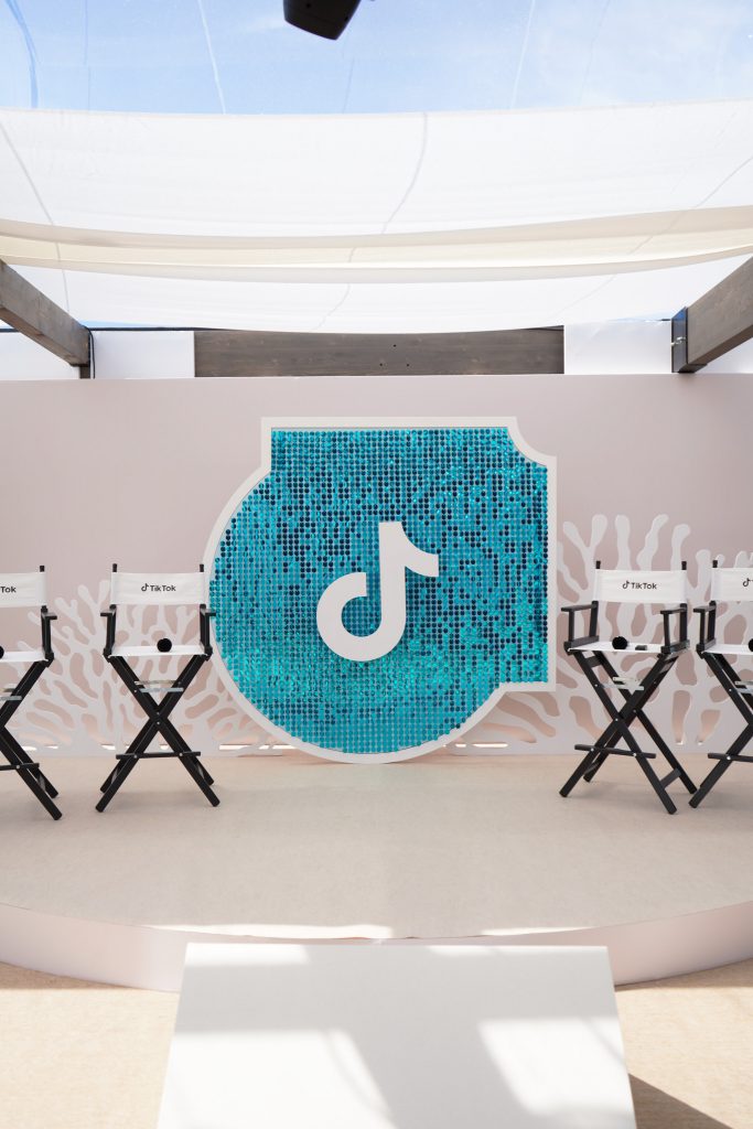 Scène sur-mesure en demi-cercle installée par AUCOP pour TikTok France à Cannes 2026 : fond de scène épuré avec motifs de coraux blancs en Forex, chaises de cinéma noires et blanches logotées et panneau central en sequins turquoise brillants.