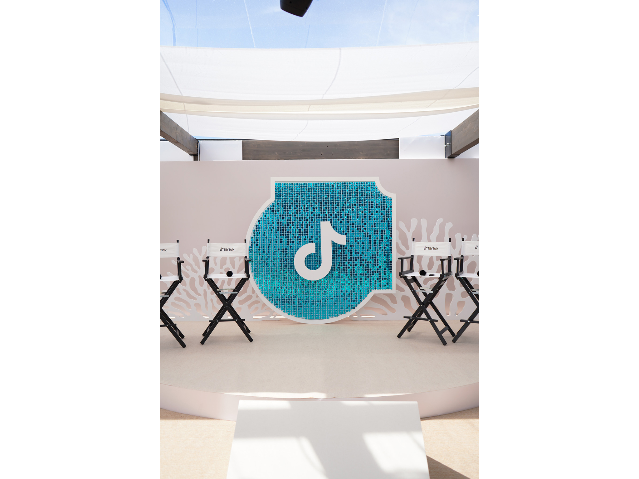 Scène sur-mesure en demi-cercle installée par AUCOP pour TikTok France à Cannes 2026 : fond de scène épuré avec motifs de coraux blancs en Forex, chaises de cinéma noires et blanches logotées et panneau central en sequins turquoise brillants.