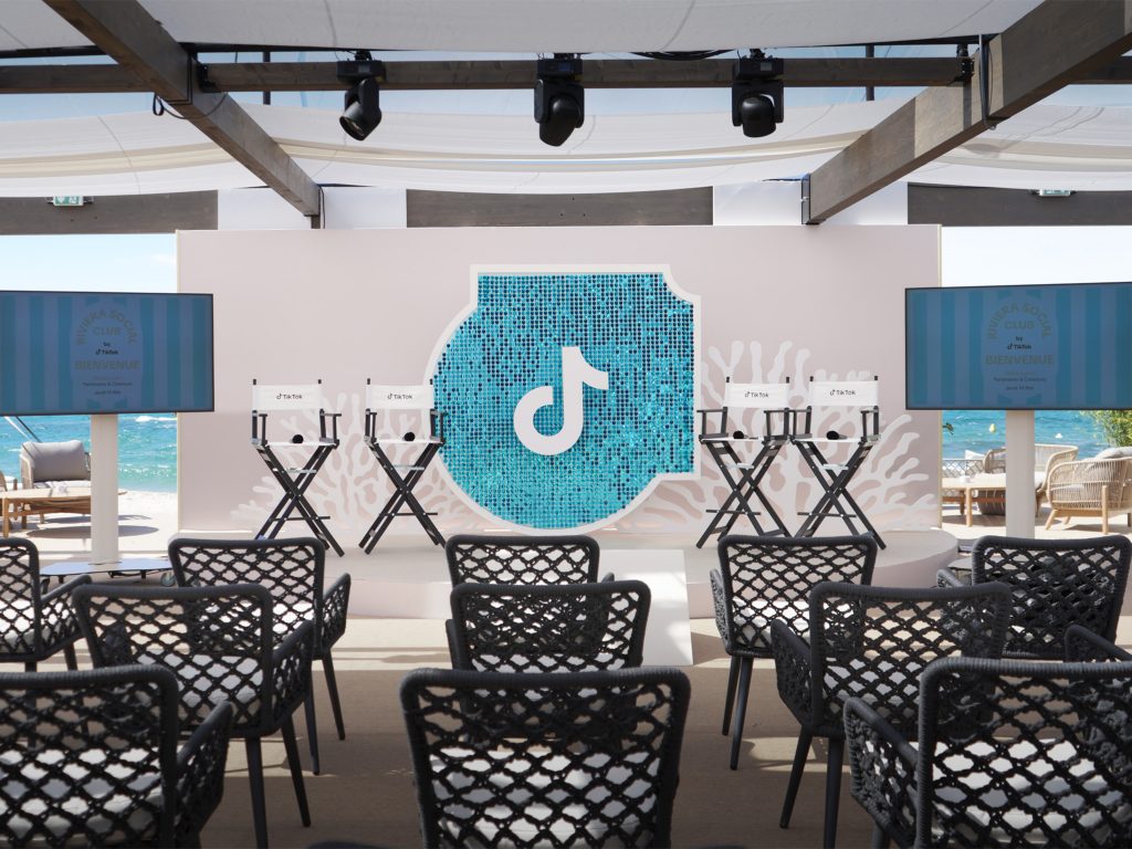 Vue d'ensemble de l'espace éphémère TikTok au Festival de Cannes 2026 sur la Plage Vegaluna conçu par AUCOP : chaises de réalisateur sur scène, panneaux de corail en Forex, grand sparkle en sequins turquoise avec le logo TikTok et écrans de diffusion sous une structure ombragée