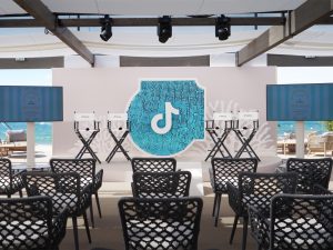 Vue d'ensemble de l'espace éphémère TikTok au Festival de Cannes 2026 sur la Plage Vegaluna conçu par AUCOP : chaises de réalisateur sur scène, panneaux de corail en Forex, grand sparkle en sequins turquoise avec le logo TikTok et écrans de diffusion sous une structure ombragée