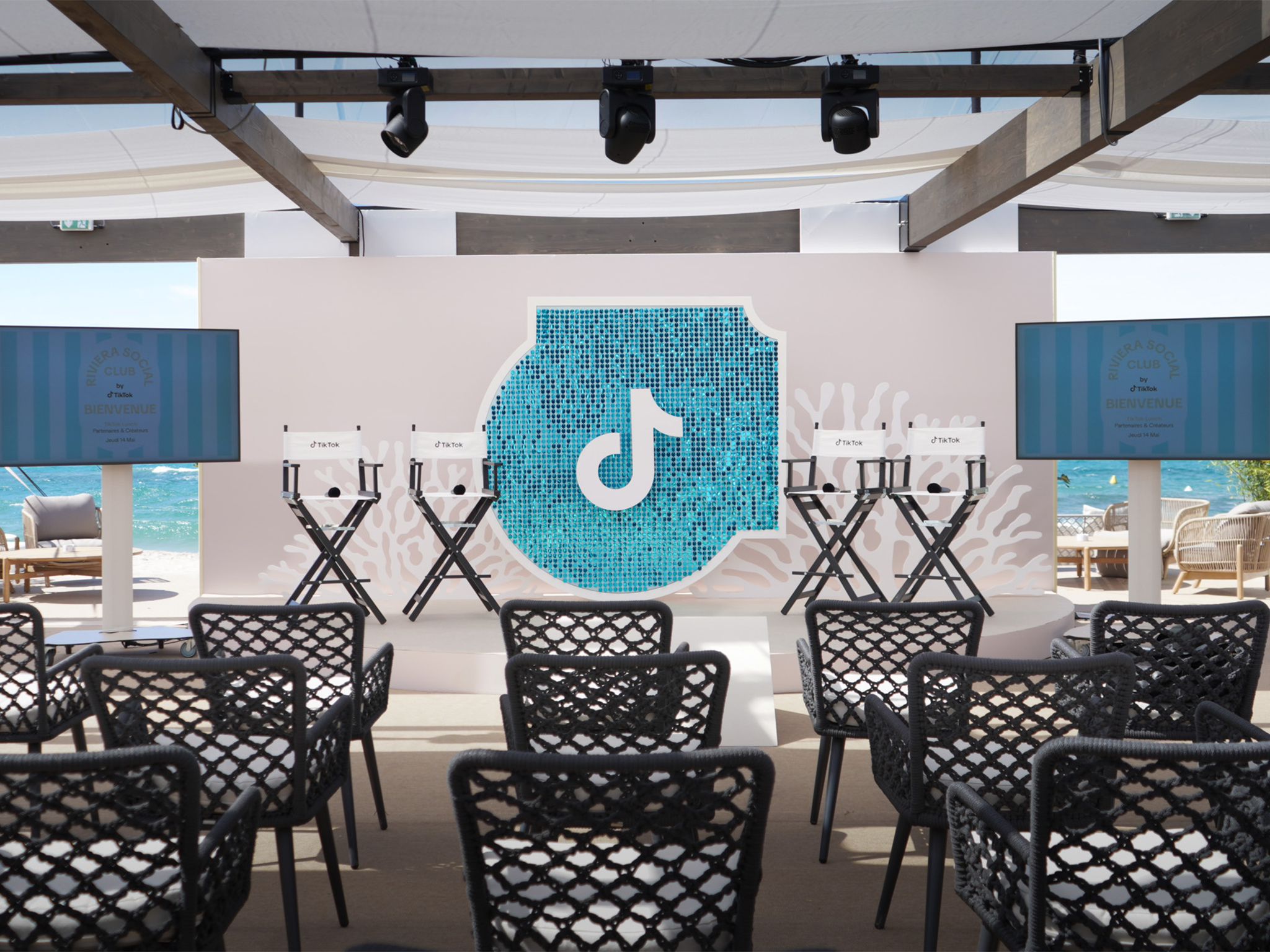 Vue d'ensemble de l'espace éphémère TikTok au Festival de Cannes 2026 sur la Plage Vegaluna conçu par AUCOP : chaises de réalisateur sur scène, panneaux de corail en Forex, grand sparkle en sequins turquoise avec le logo TikTok et écrans de diffusion sous une structure ombragée