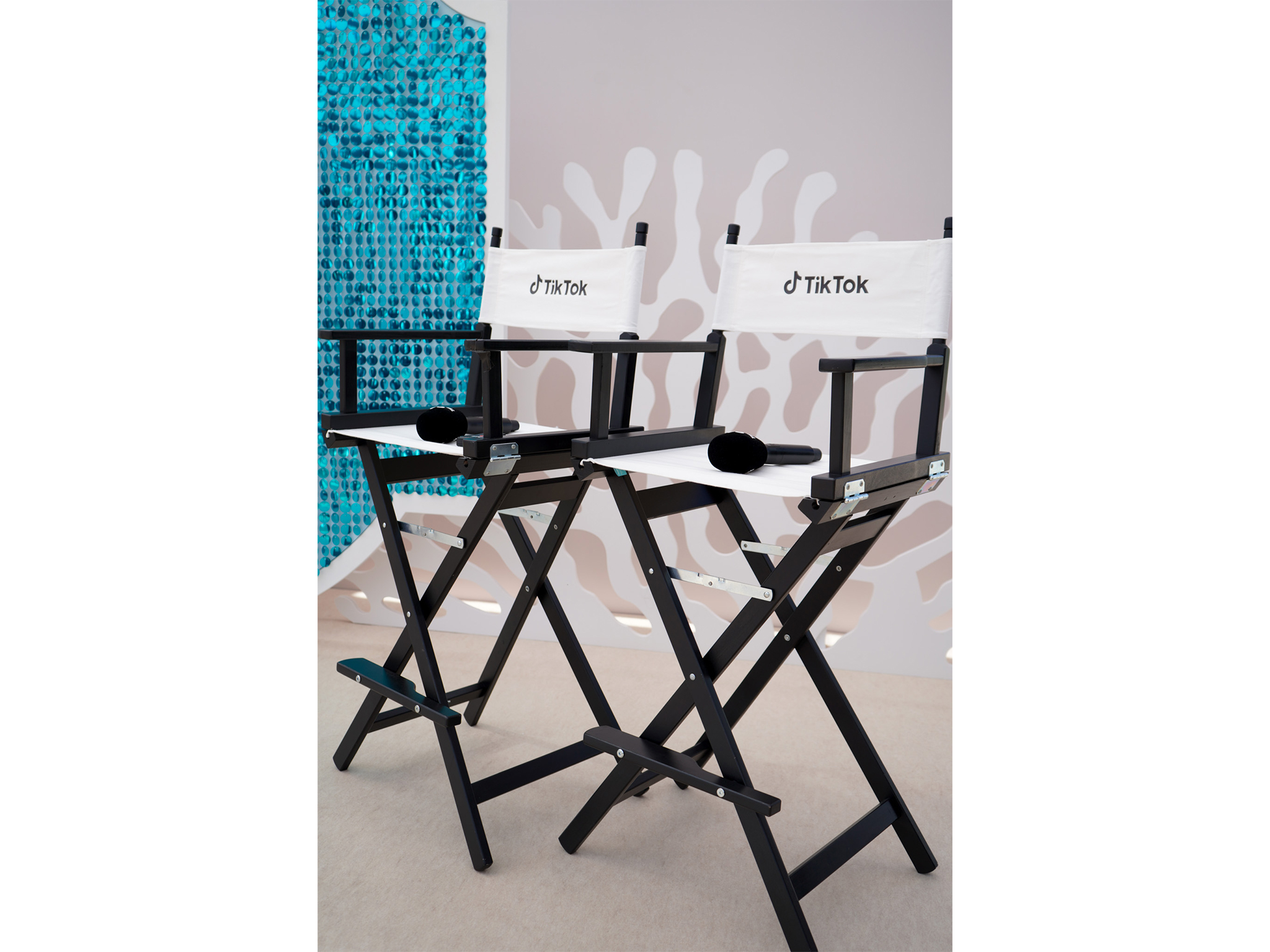 Zoom sur deux chaises de réalisateur noires et blanches personnalisées avec le logo TikTok, disposées sur la scène de la Plage Vegaluna avec des micros, devant le décor en sequins et Forex signé AUCOP.