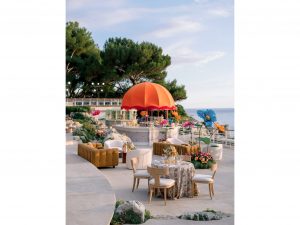 Vue d'ensemble du bar central arrondi avec dôme orangé à franges et grandes fleurs géantes décoratives, entouré de convives lors du cocktail du Welcome Dinner d'Angelina & Kelvin — production AUCOP à l'Hôtel du Cap Eden Roc, vue sur la Méditerranée.