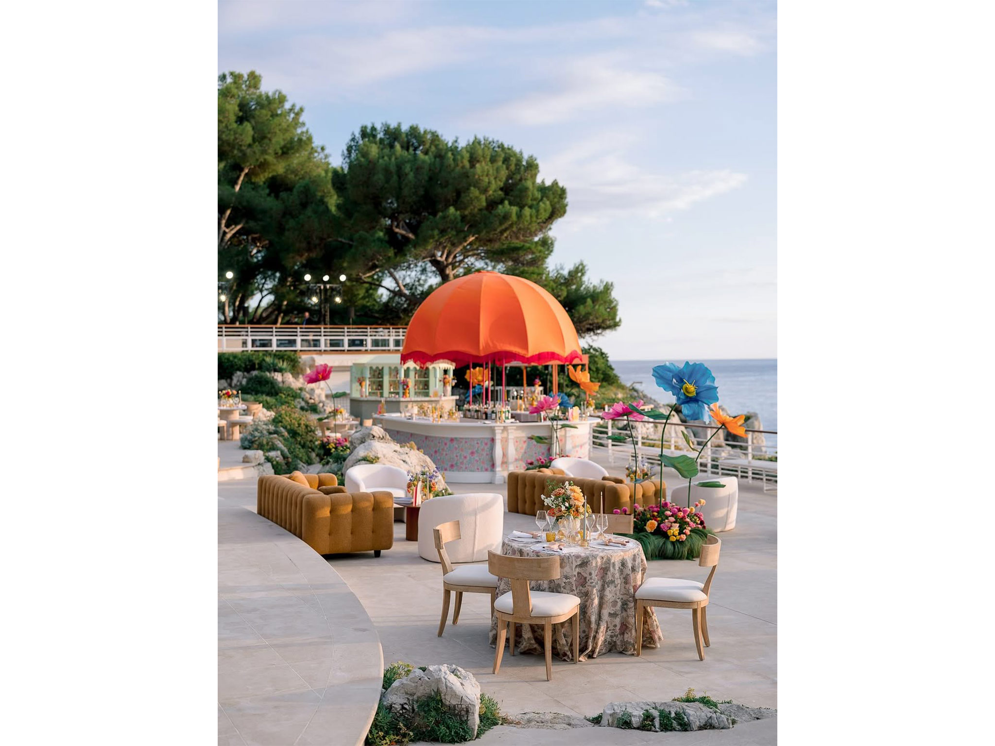 Vue d'ensemble du bar central arrondi avec dôme orangé à franges et grandes fleurs géantes décoratives, entouré de convives lors du cocktail du Welcome Dinner d'Angelina & Kelvin — production AUCOP à l'Hôtel du Cap Eden Roc, vue sur la Méditerranée.