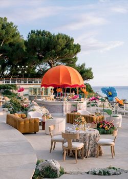Vue d'ensemble du bar central arrondi avec dôme orangé à franges et grandes fleurs géantes décoratives, entouré de convives lors du cocktail du Welcome Dinner d'Angelina & Kelvin — production AUCOP à l'Hôtel du Cap Eden Roc, vue sur la Méditerranée.
