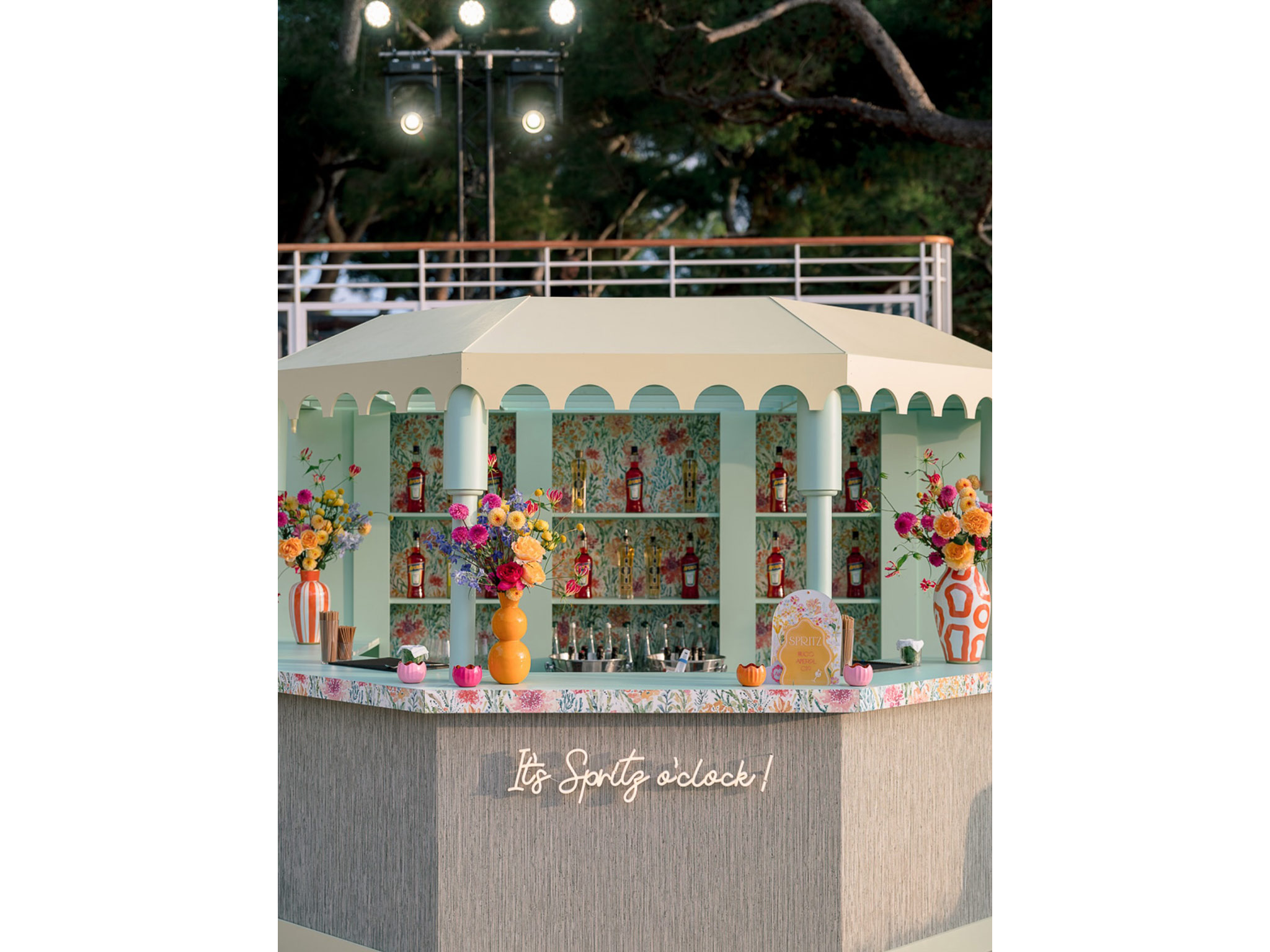 Bar à spritz octogonal sur mesure conçu et fabriqué par les ateliers AUCOP pour le Welcome Dinner d'Angelina & Kelvin à l'Hôtel du Cap Eden Roc — structure vert menthe, toit crème à festons, habillage fleuri et enseigne néon « It's Spritz o'clock ! », Antibes.