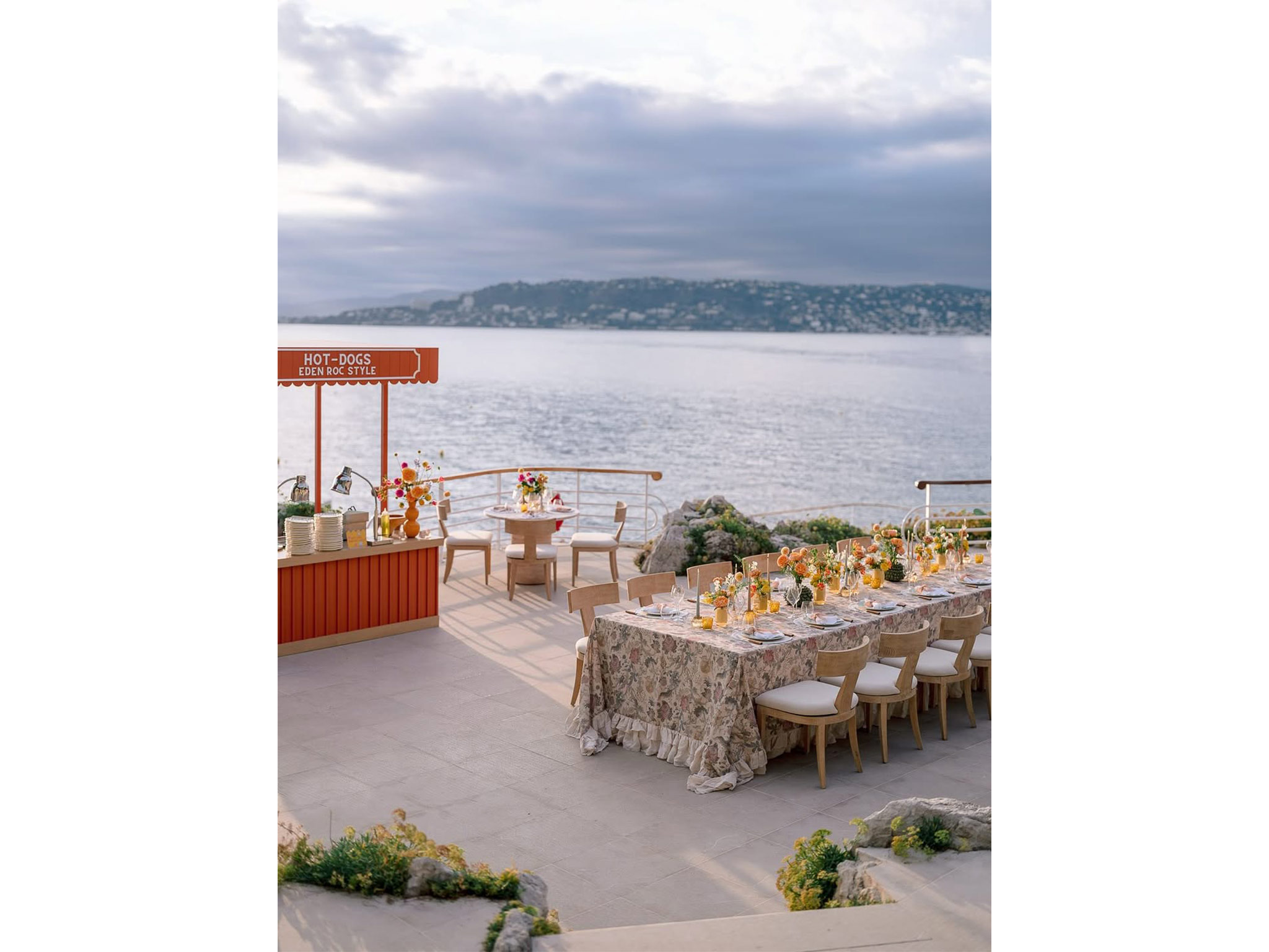 Food station « Hot-Dogs Eden Roc Style » sur mesure fabriquée par AUCOP — structure rouge à marquise, tables dressées en nappes fleuries avec verres et bougies, vue sur la Méditerranée, Welcome Dinner Angelina & Kelvin, Hôtel du Cap Eden Roc.