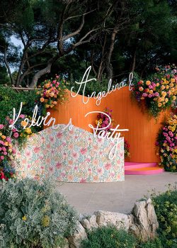 Panneau de bienvenue « Angelina & Kelvin's Party » en néon blanc sur fond orange corail, entouré de compositions florales colorées — décor fabriqué par AUCOP pour le Welcome Dinner à l'Hôtel du Cap Eden Roc, Antibes.