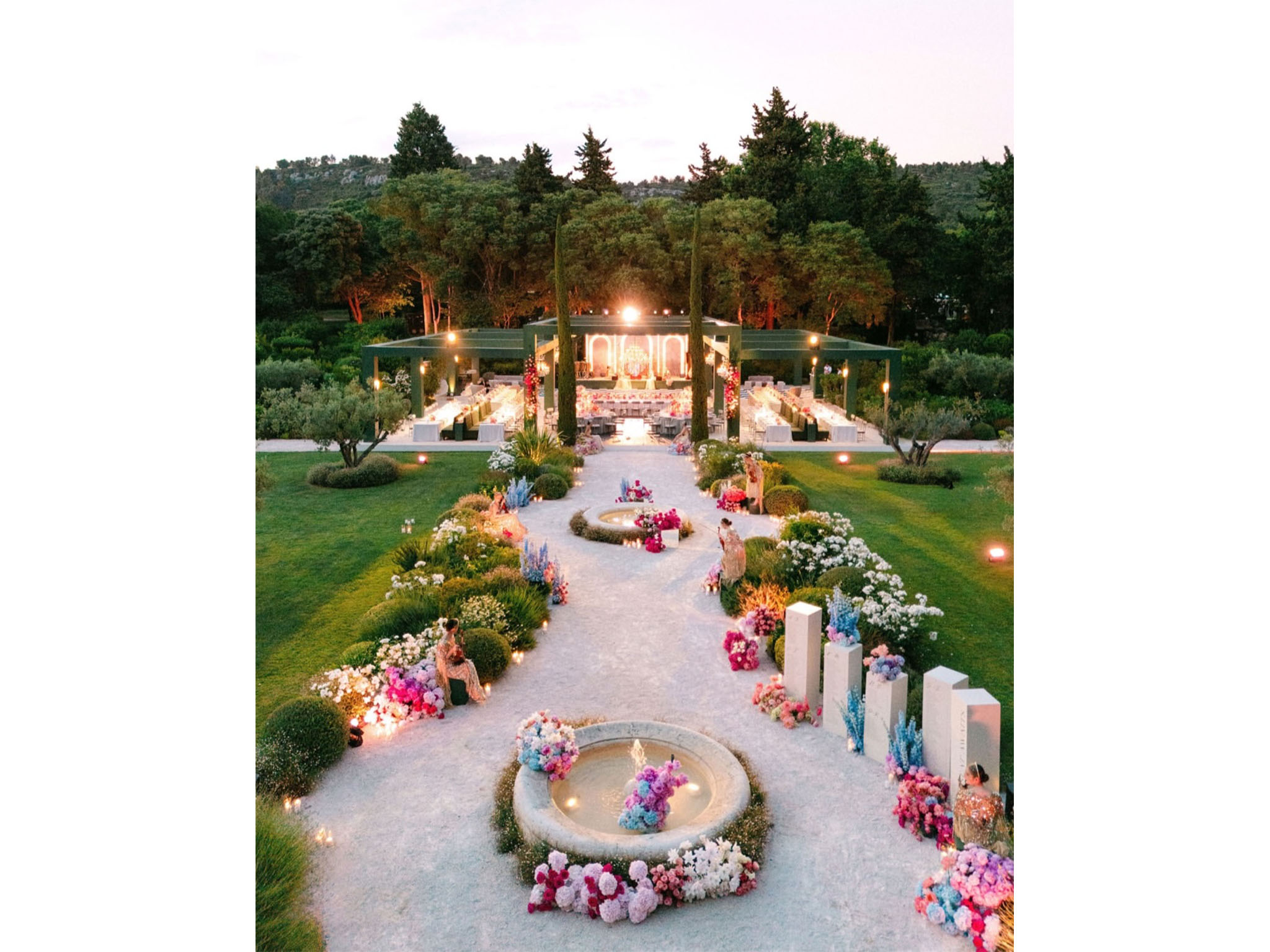 AUCOP - Vue aérienne au crépuscule du dîner de mariage : pergola-structure verte illuminée, allée en gravier avec fontaines fleuries et stèles florales, jardin provençal - scénographie White Eden Weddings, production AUCOP, Airelles Château d'Estoublon, Provence