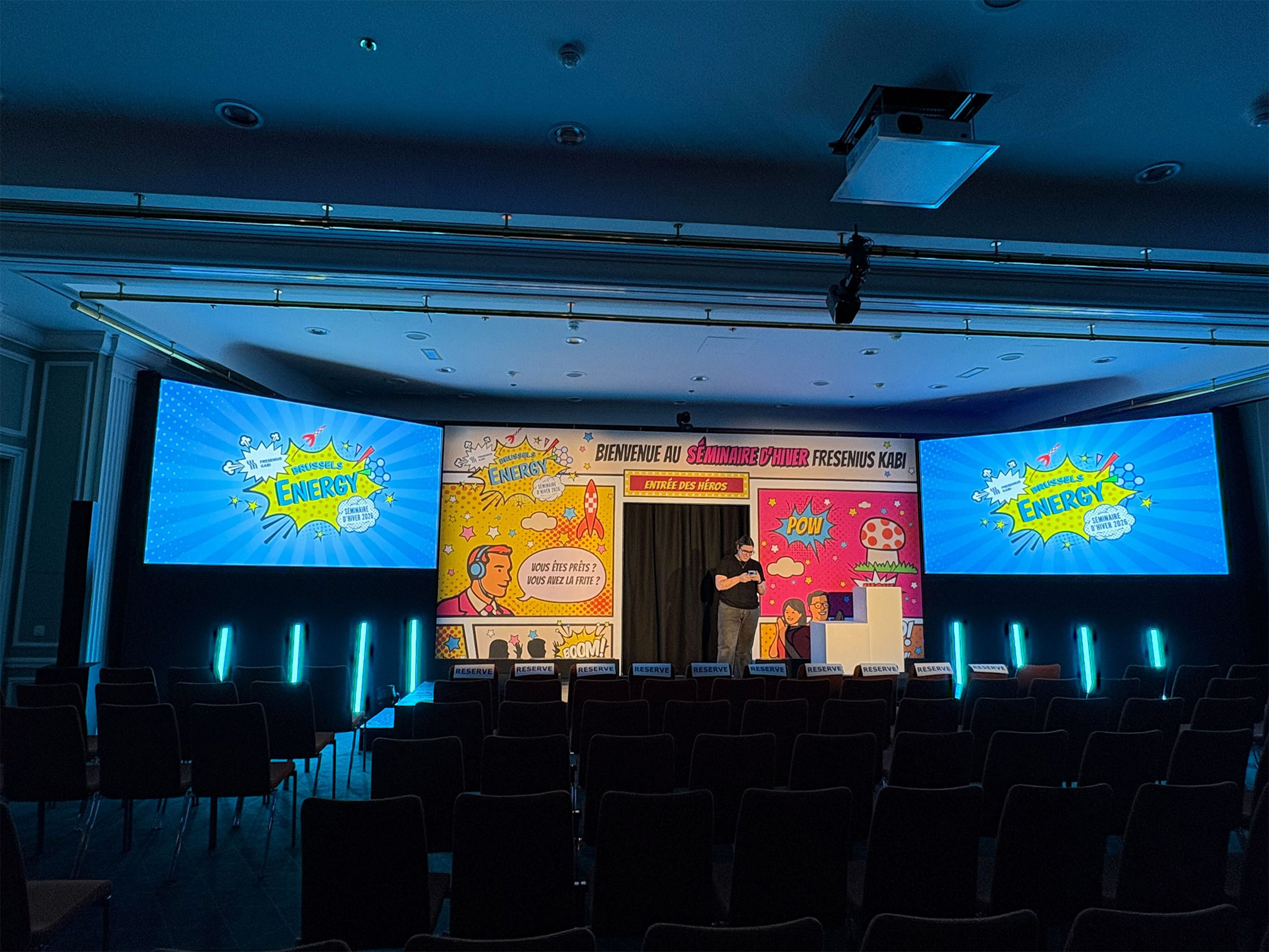 QUAND LA SCENOGRAPHIE DEVIENT UNE BANDE DESSINEE GEANTE Salle du séminaire d'hiver Brussels Energy de Fresenius Kabi au Hilton de Bruxelles — fond de scène bande dessinée grand format, écrans latéraux et colonnes LED cyan, réalisé par Aucop Paris pour l'agence Enkorprod